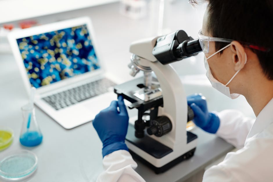 Scientist examining samples under a microscope in a lab with laptop display.
